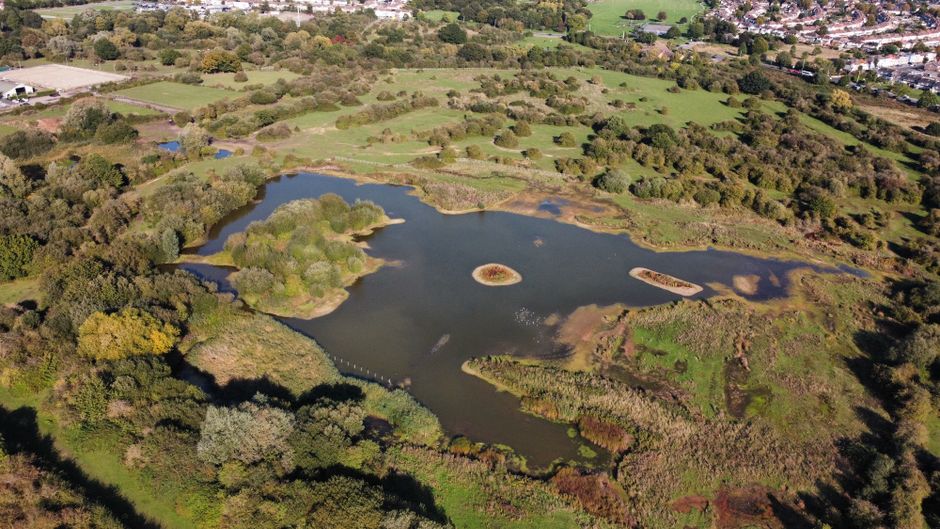 An aerial view of a wetland area near Barnes, showing a large pond with an irregular shape, small islands, and surrounding greenery. The pond features a central circular island and smaller patches of 